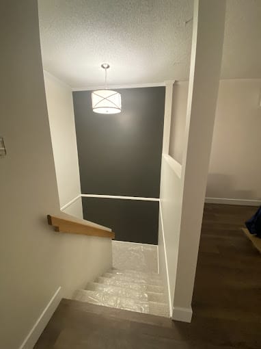 A well-lit staircase with wooden handrails descends from a white-walled hallway in this new home. The stairs lead to a lower level with dark wooden flooring, while a modern, circular light fixture hangs from the ceiling, casting light on the staircase and walls.