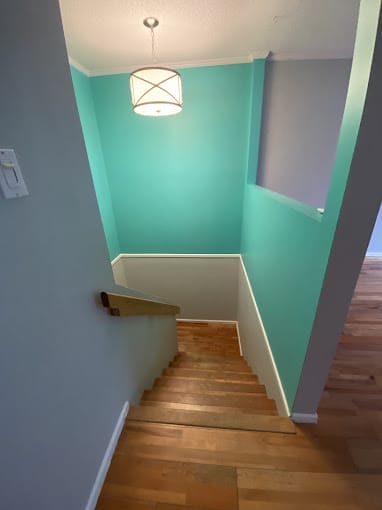 A staircase descends with wooden steps, enclosed by light blue walls and white trim. At the bottom, where the new light grey walls take over, a modern ceiling light hangs above, illuminating the area elegantly. There is a visible hardwood floor at the bottom of this inviting home feature.