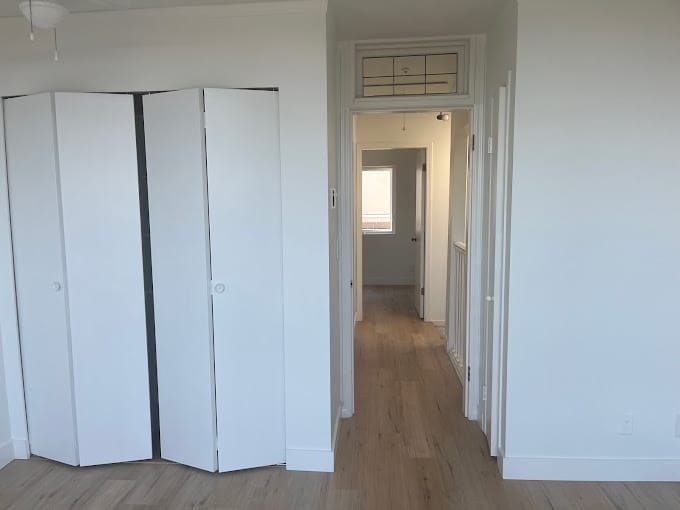 A brightly lit hallway in a new, modern home with white walls and light wooden flooring. A closed white closet door is on the left side. Above the door frame, there is a decorative glass windowpane. Sunlight streams in from a window at the end of the hallway, illuminating every page of this beautiful space.