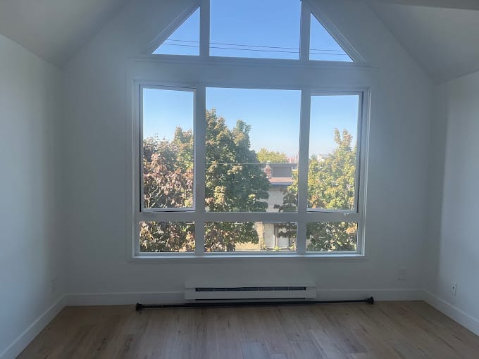 A bright room with a large, triangular set of windows showcasing a view of trees and a clear blue sky outside. The room has light-colored wooden flooring and white walls, creating an airy and spacious atmosphere—the perfect inspiration for your New Home Page.